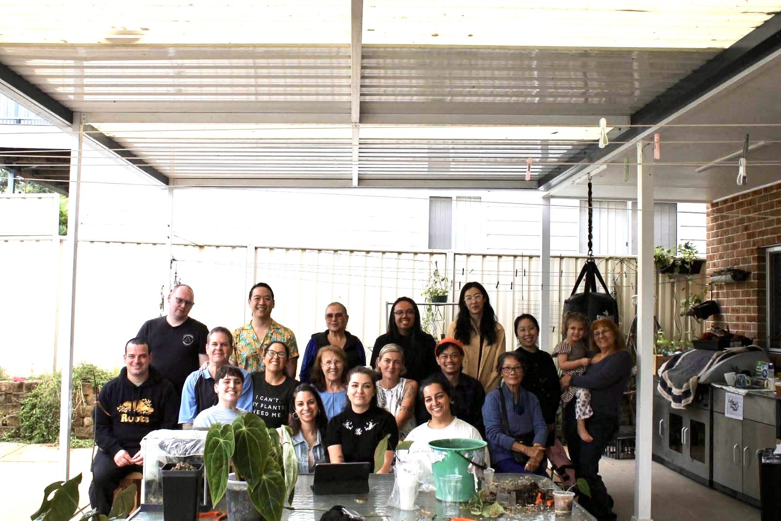 About twenty people seated and standing together under a pergola, with plants and a potting table in front of them.